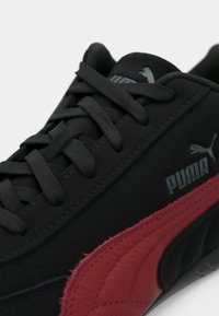 Fekete sneaker, texturált felső résszel, szürke Puma márkázással és piros bőr oldalcsíkkal. A fűzők feketék, az összhatás matt.