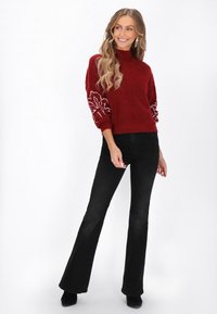 Czerwony sweter z wysokim kołnierzem i teksturowanym materiałem, z białą haftowaną florystyką na rękawach. Połączony z czarnymi spodniami dzwonami.