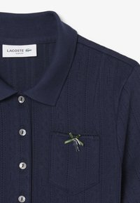 Marineblå polo-shirt med et struktureret mønster, ribbet krave, sølvknapper og en brystlomme med et grønt bånd og sølvdetaljer.