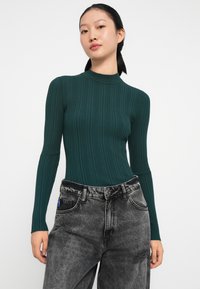 Grünes, geripptes Langarmoberteil mit hohem Ausschnitt, kombiniert mit hochtaillierten, ausgewaschenen schwarzen Jeans, die über Taschen vorne und leichte Abnutzungen verfügen.