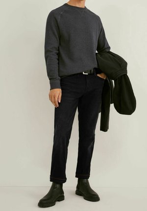 Grau Crewneck-Pullover, schwarze Jeans, grüne Stiefeletten. Schwarze Jacke über dem Arm drapiert. Schmaler Schnitt und minimalistisches Design.