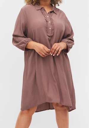 Robe ample mauve à manches longues, patte de boutonnage à volants et ourlet arrière légèrement plus long, portée par une personne les mains jointes.