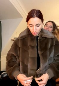 Person mit roten Haaren trägt eine braune Pelzjacke und schließt den Reißverschluss, eine weitere Person mit langen Haaren steht dahinter in einem Raum mit schräger Decke.