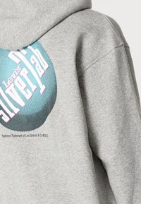 Sudadera de color gris claro con un gran diseño gráfico en color verde azulado y blanco, con detalles en rosa. El diseño incluye el texto "Levi's Silverlab" en una fuente negrita.