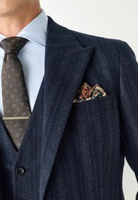 Tengerész kék csíkos zakó, kis csúccsal, barna mintás nyakkendővel és vörös, arany és tengerész kék színű paisley zsebkendővel párosítva.