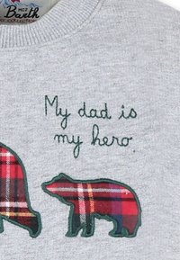 Felpa grigia con testo ricamato "Mio padre è il mio eroe", affiancata da due forme di orso decorate con motivi a quadretti rossi e neri.