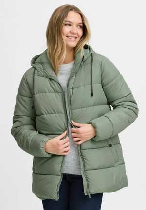Doudoune verte avec capuche, design matelassé, fermeture éclair et cordons noirs. Portée par-dessus un pull gris, avec des poignets élastiques.