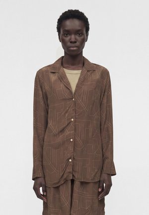 Άτομο με κοντά σγουρά μαλλιά φοράει καφέ πουκάμισο με κουμπιά και ταιριαστό παντελόνι με ανοιχτά αφηρημένα γραμμικά μοτίβα πάνω από μπεζ τοπ.