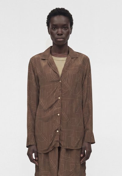 Personne aux cheveux courts et bouclés porte une chemise marron à boutons et un pantalon assorti avec des motifs de lignes abstraites claires sur un haut beige.