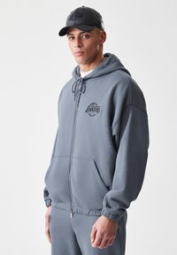 Felpa grigia con zip, tasche a marsupio e polsini elasticizzati, con il logo nero dei Los Angeles Lakers sul petto. Tessuto dalla texture liscia.
