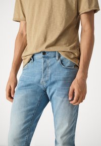 Hombre con vaqueros de mezclilla azul claro y camisa de manga corta beige, de pie con los brazos relajados a los lados.