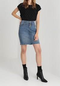 Top negro texturizado, falda de mezclilla azul de tiro alto con cierre de botones en el frente y botas negras hasta el tobillo con puntas afiladas y tacones de aguja.
