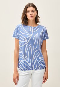 Haut à manches courtes bleu en tissu doux, avec des motifs de feuilles blanches, un col rond et une coupe décontractée. Porté avec un pantalon blanc.