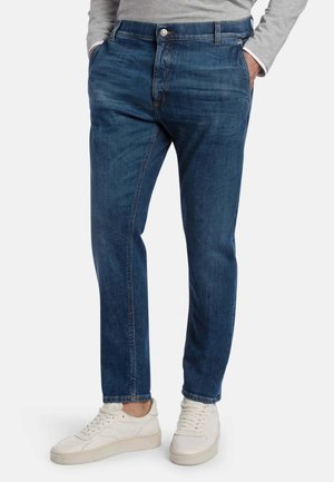 Uomo che indossa jeans blu slim fit, camicia grigia a maniche lunghe e sneakers bianche, con le mani in tasca, in piedi su sfondo bianco.