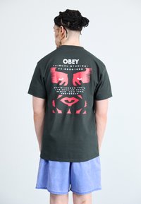 Black cotton t-shirt featuring red graphic design, "OBEY" text, and stylized faces on the back. Paired with light blue shorts.