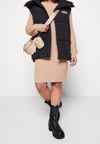 Doudoune noire avec capuche, robe pull beige côtelée, et bottes noires à talons épais. Sac bandoulière beige avec chaîne.