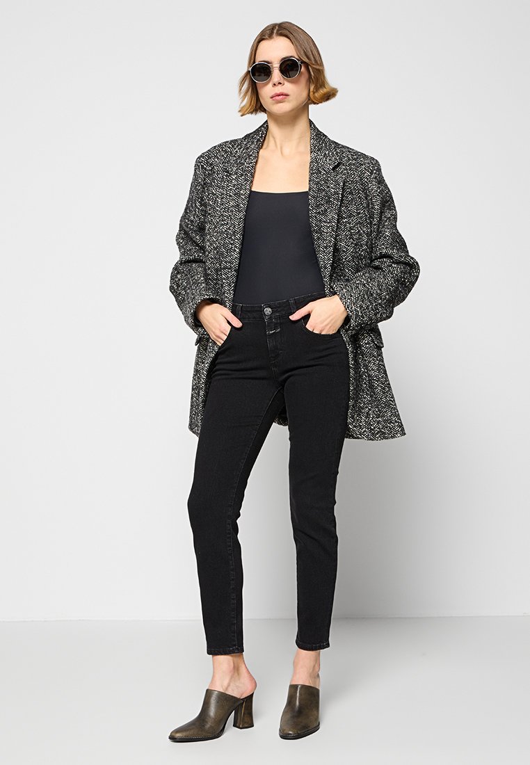 Schwarzer und weißer Hahnentrittmantel mit Oversize-Passform, kombiniert mit einem enganliegenden schwarzen Oberteil, schwarzen Skinny-Jeans und dunklen, hohen Mules.