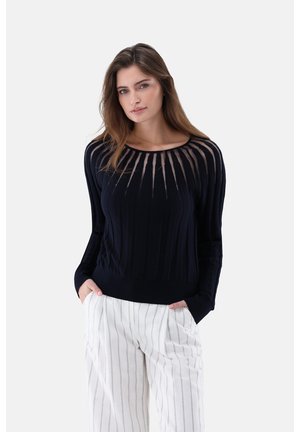 Femme portant un pull noir avec des rayures verticales transparentes au niveau de l'encolure et un pantalon blanc à fines rayures verticales, les mains dans les poches.