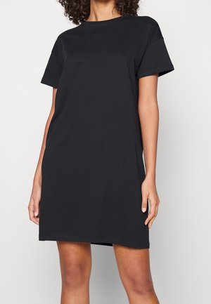 Osoba nosząca prostą czarną sukienkę w stylu t-shirt z krótkim rękawem i okrągłym dekoltem na jednolitym białym tle.