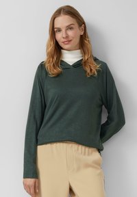 Grüner Kapuzenpullover mit weicher Textur, entspannter Passform und langen Ärmeln; getragen über einem weißen Rollkragenpullover mit hellbeigen Hosen.