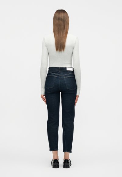 Γυναίκα που φαίνεται από πίσω, φοράει ανοιχτό γκρι μπλούζα με μακριά μανίκια, σκούρα μπλε τζιν ψηλόμεσο και μαύρα μοκασίνια, σε ένα απλό φόντο.