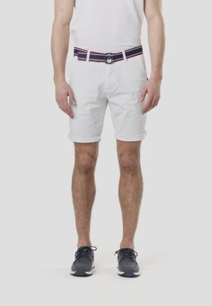 Białe bawełniane szorty z podwiniętymi mankietami, z granatowym paskiem w czerwone i białe paski. Model ma na sobie granatowe trampki.