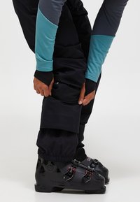 Svarta skidbyxor med elastiska muddar, tillsammans med svarta skidstövlar med röda och silverfärgade spännen. Överdelens ärmar är blå och grå.