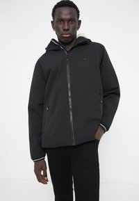 Jachetă neagră cu glugă, cu închidere cu fermoar, buzunare laterale și garnituri contrastante pe mansete. Căptușeală moale vizibilă în interiorul glugii.