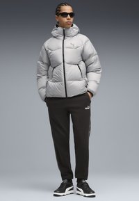 Jachetă puffer argintie cu guler înalt și fermoar, purtată cu pantaloni de trening negri și adidași negri cu talpă albă și logo.