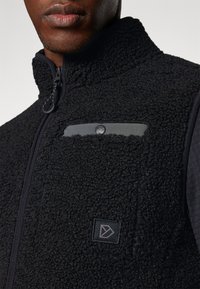 Μαύρο γιλέκο από φλις με υφασμάτινη υφή, με φερμουάρ και μία μικρή γκρι τσέπη με κουμπί στην επάνω αριστερή πλευρά.