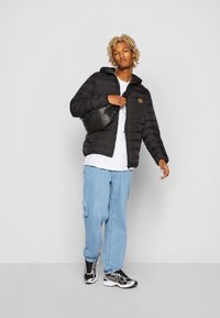 Sort puffjakke med hætte, lynlås lukning og et beige logo patch. Kombination med en hvid t-shirt, lyseblå jeans og sorte sneakers.