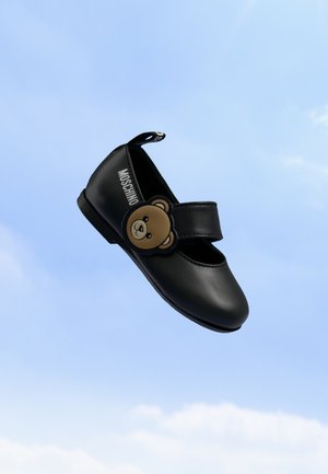Chaussure Mary Jane noire avec décoration ours en peluche et logo MOSCHINO sur le côté, sur un fond de ciel bleu clair.