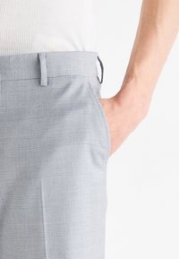 Pantaloni a quadri grigi con una texture liscia, dotati di vita piatta, tasche laterali e una cintura aderente, mostrati con una mano appoggiata in tasca.