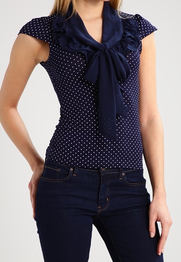 Mörkblå blus med prickar, korta ärmar och en volangkrage som avslutas med en knytning. Bärs tillsammans med mörka jeans, vilket framhäver den figursydda designen.