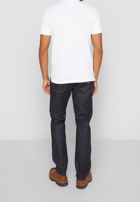 T-shirt blanc à manches courtes, jean bleu foncé, chaussures brunes ; figure debout vue de dos. Lignes épurées, design décontracté.