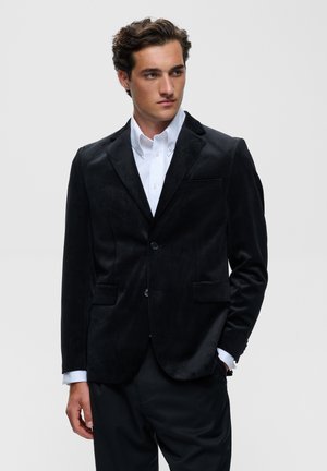 Jeune homme aux cheveux bruns ondulés porte un blazer en velours noir sur une chemise blanche boutonnée, regardant vers la droite avec une main dans sa poche.