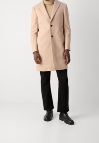 Abrigo de lana beige con un corte ajustado, diseño de un solo botón, solapas tipo muesca y dos bolsillos frontales, combinado con pantalones negros y botas.