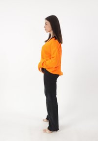 Maglione arancione oversize a maglia con polsini a coste, abbinato a jeans neri aderenti e sandali con tacco beige, visto di lato.