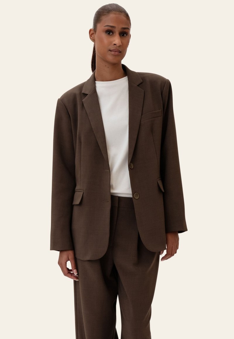 Femme aux cheveux tirés en arrière portant un blazer marron foncé ample et un pantalon assorti sur un t-shirt blanc à col rond, debout devant un fond clair.