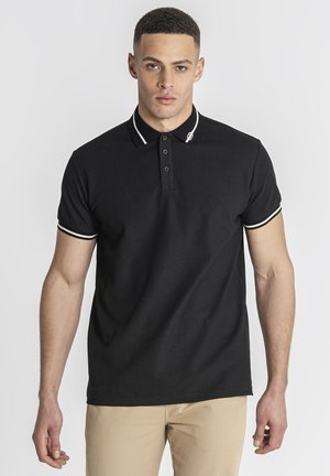 Man draagt een aansluitend zwart poloshirt met witte bies aan kraag en mouwen, gecombineerd met beige broek, staand tegen een effen achtergrond.