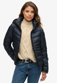 Navy pufferjack in een gewatteerd design met capuchon. Gedragen over een crèmekleurige kabeltrui en blauwe denim jeans, met een zilveren gesp riem.