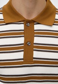 Gros plan d'un polo rayé marron, blanc et noir avec un col marron et deux boutons sur la patte de boutonnage.