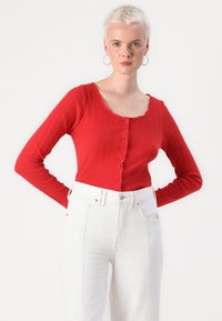 Röd ribbad långärmad topp med rundad halsringning, knäppning och vågkantad nederkant. Kombination med vita högmidjade jeans.
