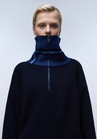 Marineblauwe pullover met hoge kraag en rits, met ribbelstructuur en contrasterende blauwe strepen op de kraag.