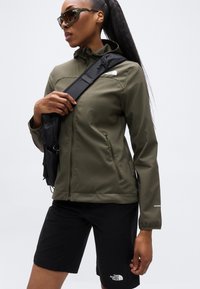 Χακί πράσινο μπουφάν με κουκούλα και τσέπες με φερμουάρ, συνδυασμένο με μαύρο σορτς. Περιλαμβάνει μαύρη τσάντα ώμου και γυαλιά ηλίου.