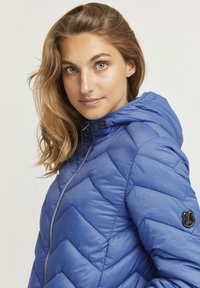 Veste isolante bleue avec capuche, présentant un motif en chevron, une fermeture à glissière et un patch logo noir rond sur la manche.