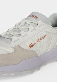Baskets blanches Lacoste avec accents en daim beige, panneau latéral matelassé, design à lacets et logo Lacoste orange avec broderie crocodile.