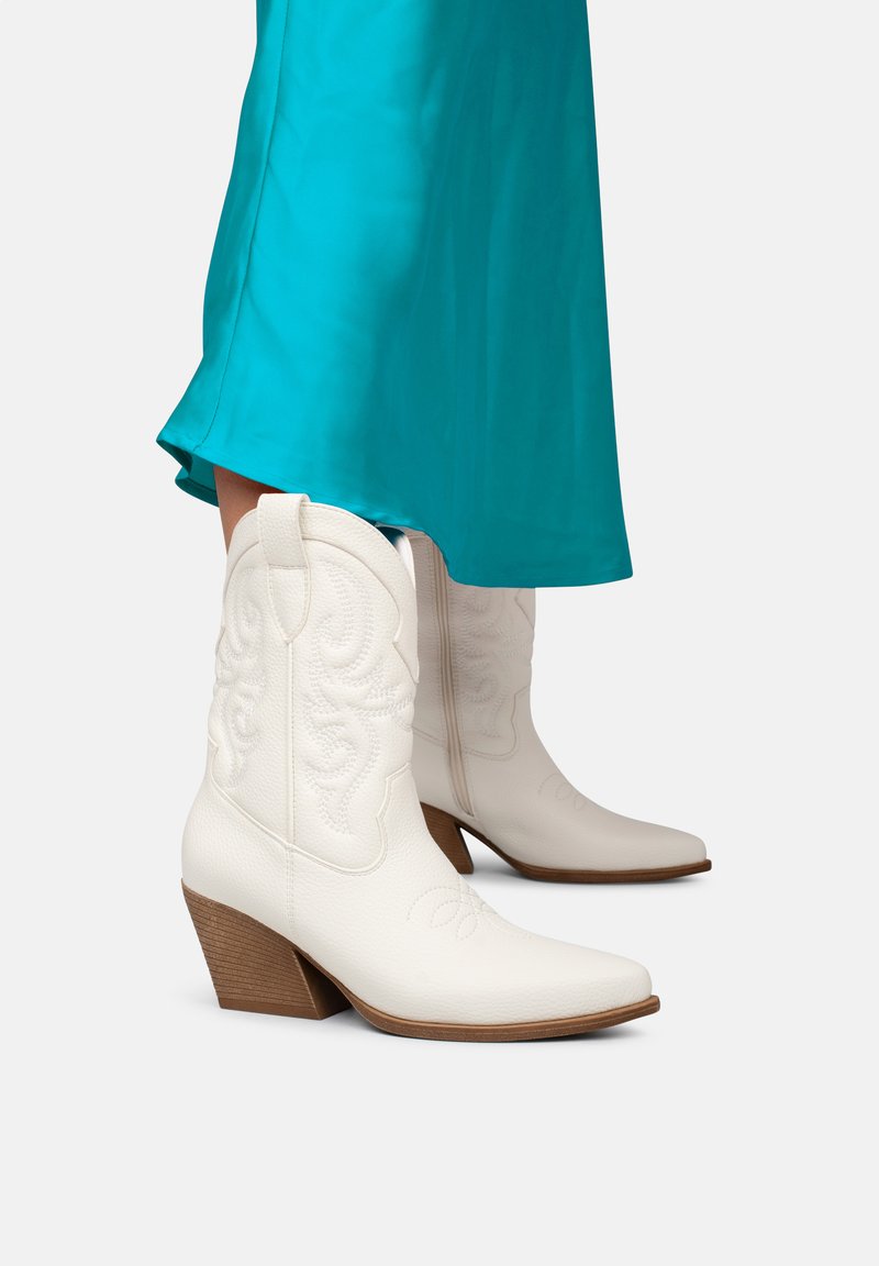 Botas de vaquero de cuero blanco con puntas afiladas, detalles en relieve y tacones altos marrones. Llevadas con una falda verde azulado visible en el dobladillo.