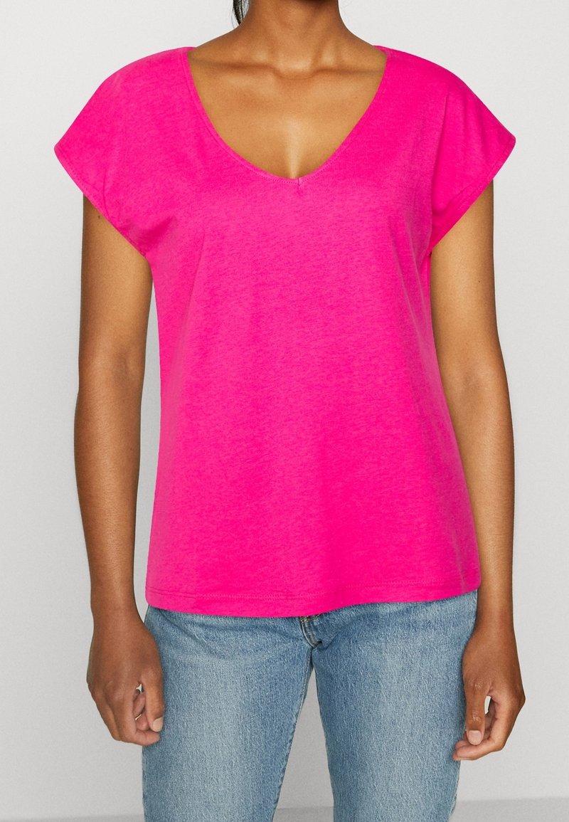Femme portant un haut rose vif à manches courtes et col en V, associé à un jean en denim bleu clair sur un fond uni.