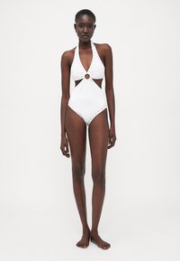 Costume da bagno intero bianco con scollatura a halter, lati con aperture e dettaglio in hardware circolare. Presenta accenti borchiati lungo i bordi.